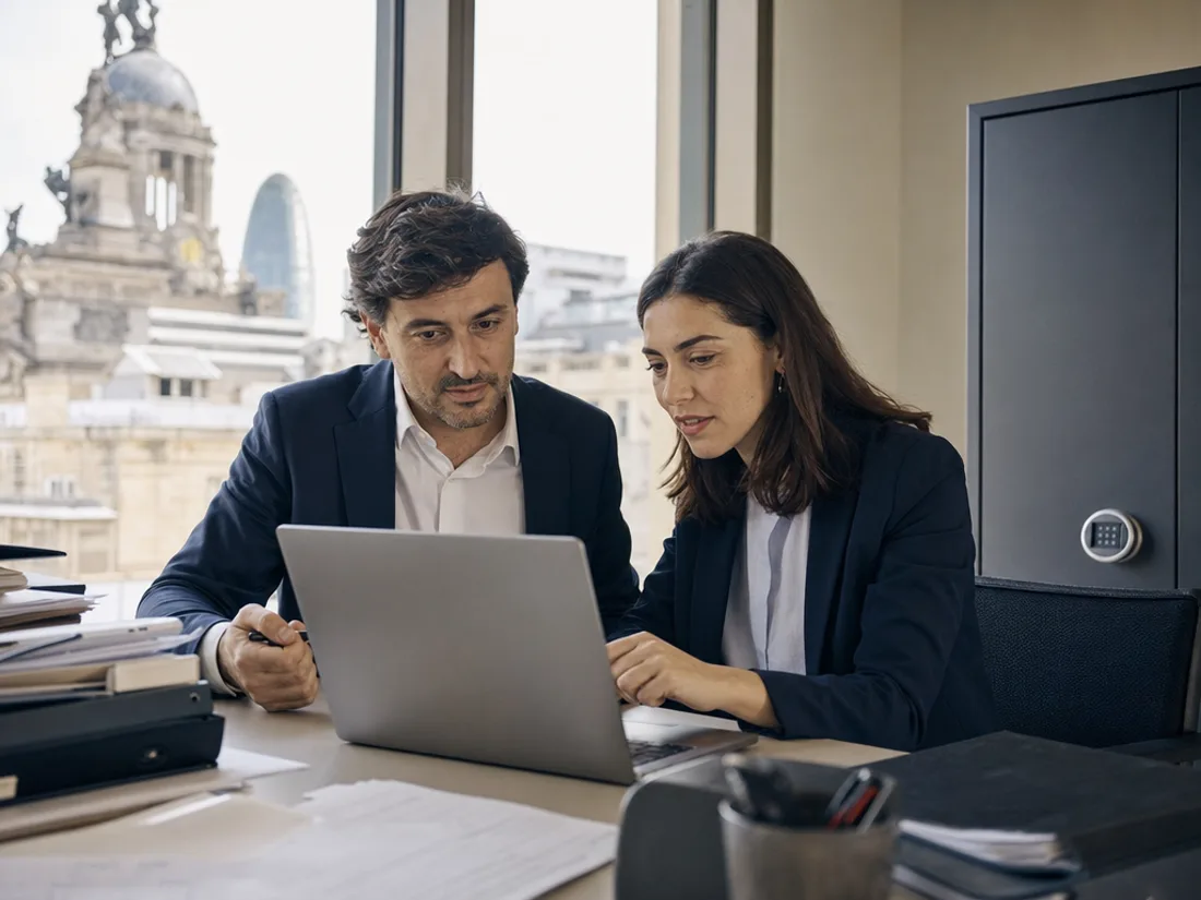 Dos profesionales revisan documentación en un despacho, con archivadores y una caja fuerte al fondo.